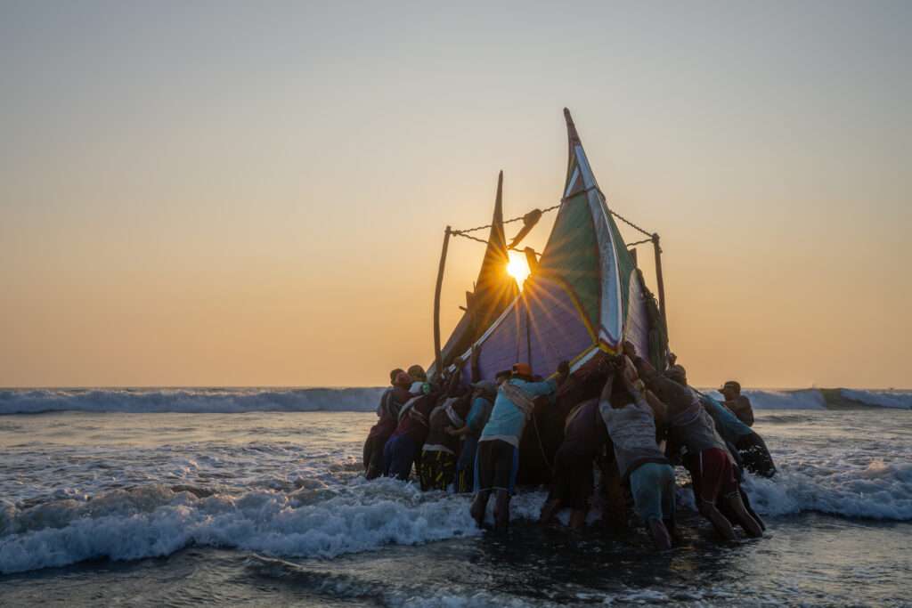 Gruppe von rund 20 Fischern schiebt bei Sonnenuntergang ein traditionelles Fischerboot ins Meer in Bangladesch